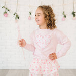 Young girl wearing a pink outfit with a heart design, standing against a light-colored wall with decorative elements.