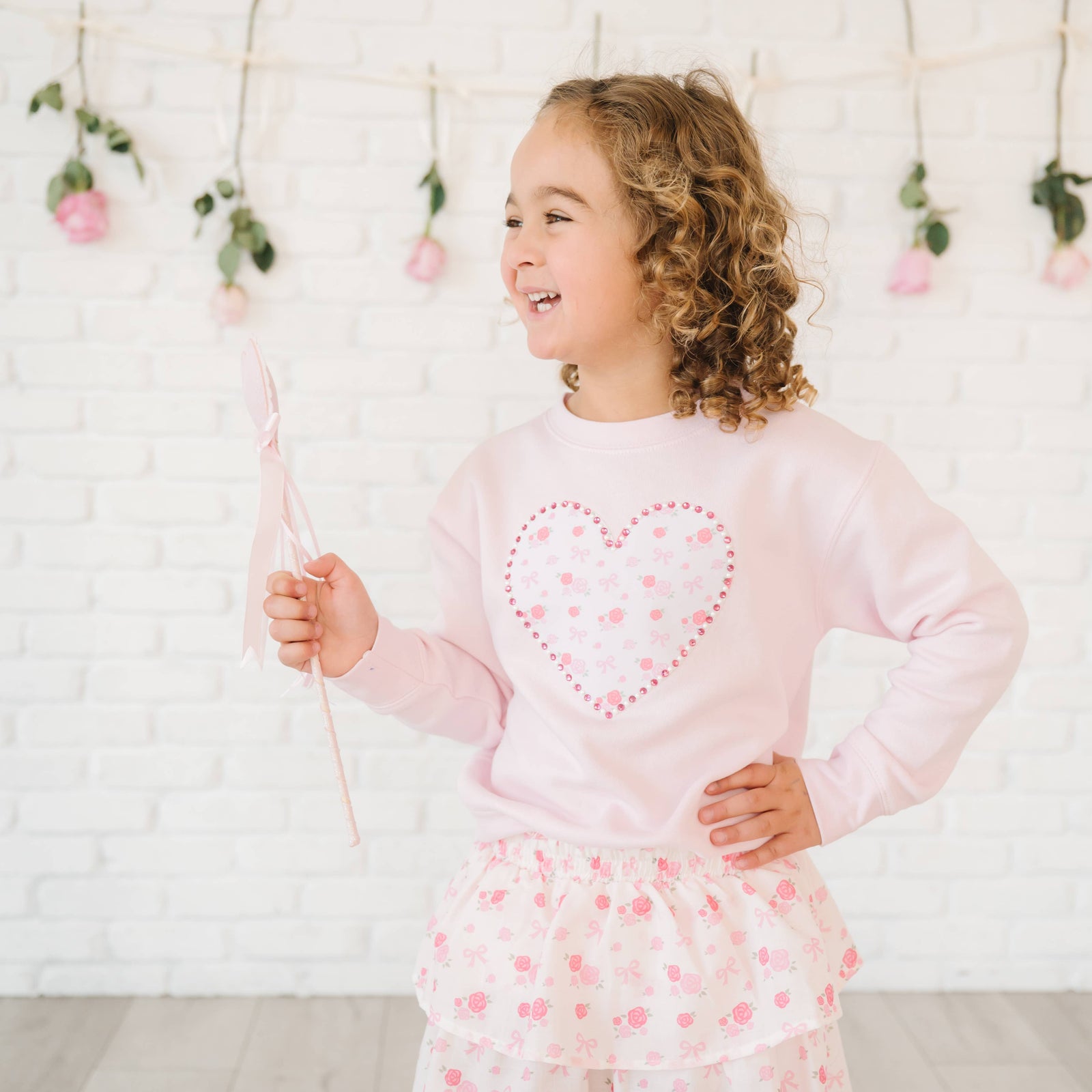 Young girl wearing a pink outfit with a heart design, standing against a light-colored wall with decorative elements.